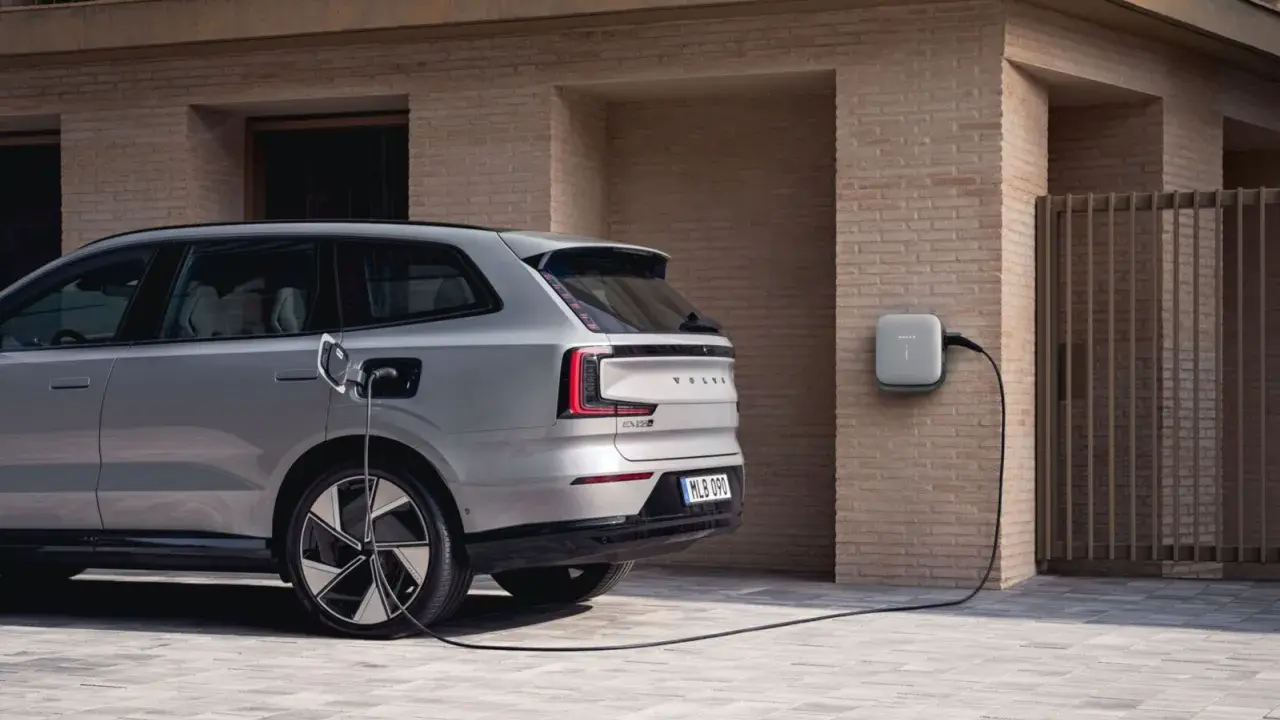 Srebrny, duży SUV Volvo EX90 podłączony do ładowarki ściennej. Nowoczesny design i ekologiczna energia.