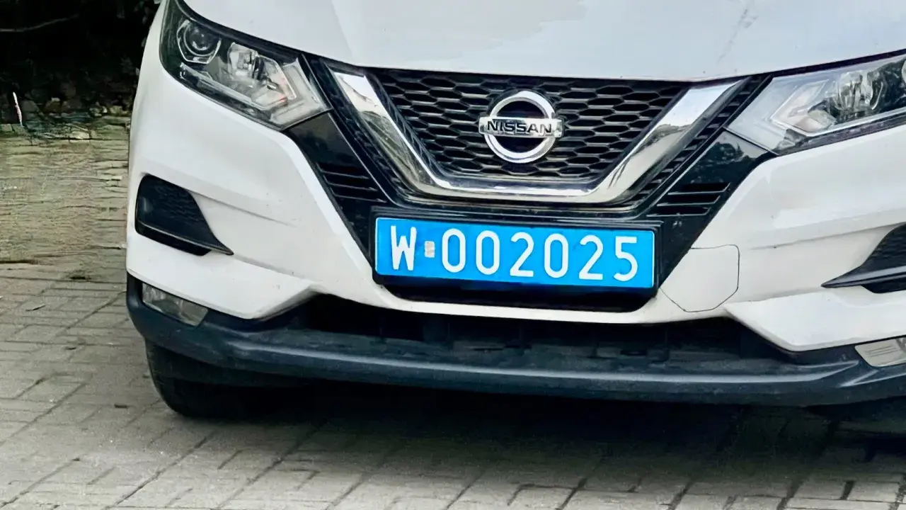 Biały Nissan z niebieską tablicą rejestracyjną W-002025, wskazującą na dyplomatyczne tablice rejestracyjne.