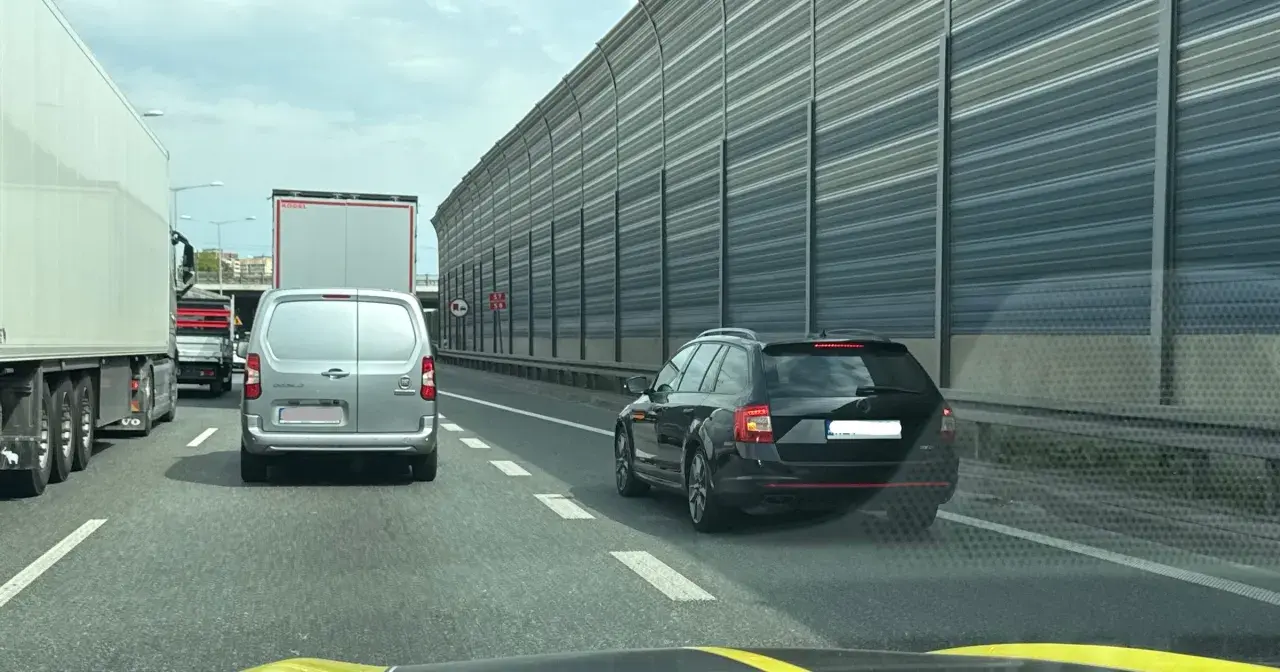 Korek na autostradzie. Samochody osobowe i ciężarowe stoją w rzędzie. W tle widać ekrany akustyczne. Jazda na suwak przepis to podstawa.