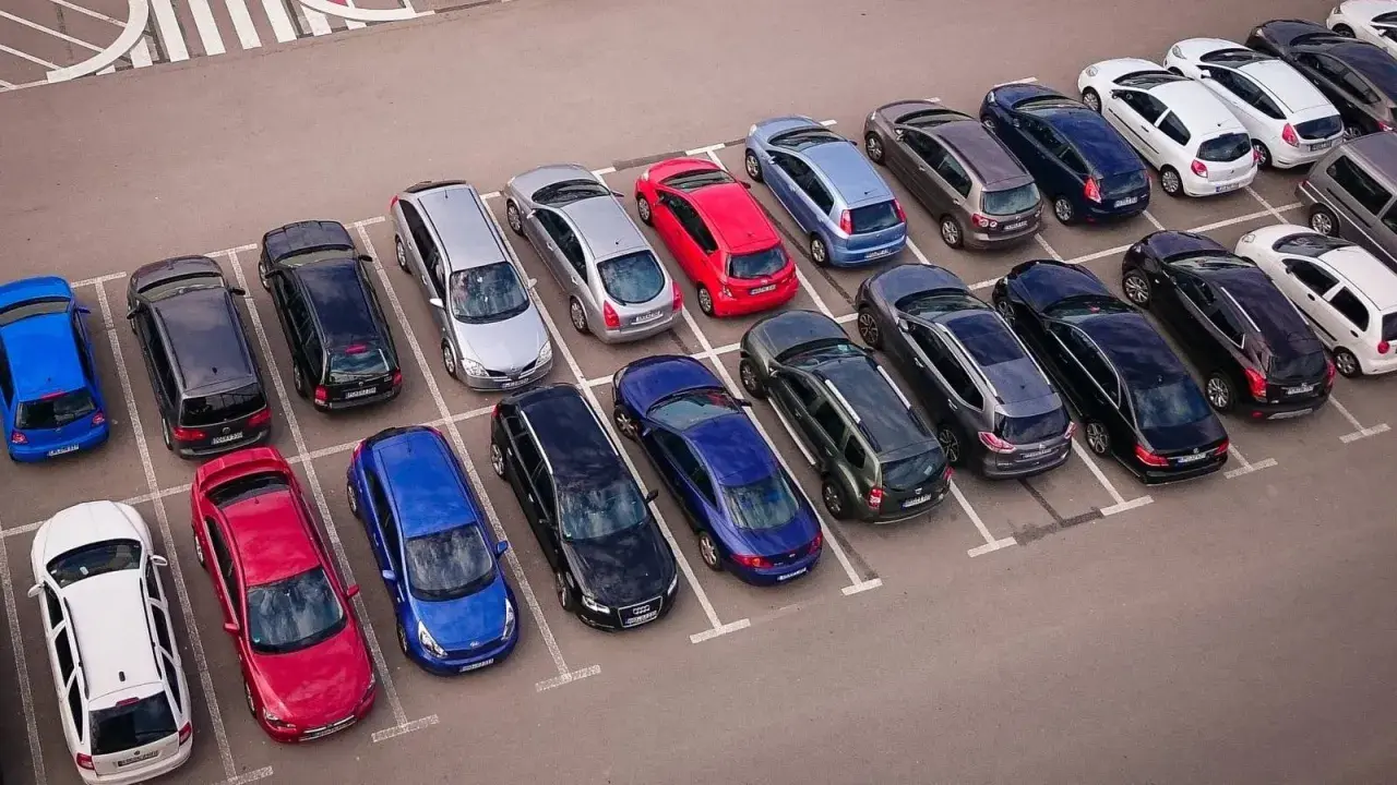 Rząd samochodów na parkingu, w tym czerwony, niebieski i srebrny. Jeden z nich czeka na odholowanie.