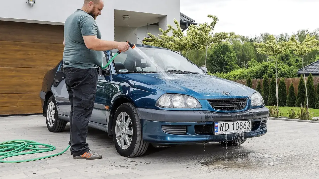 Mężczyzna myje niebieskie auto pod domem w upalny dzień. Wąż ogrodowy leży na podjeździe.