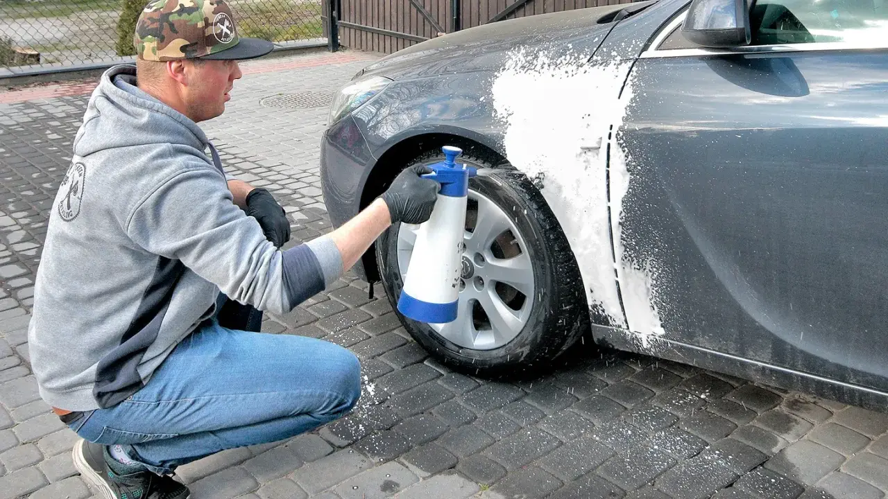 Mężczyzna w czapce z daszkiem i jeansach kuca przy samochodzie, rozpylając pianę na błotnik. Czy można naprawiać auto pod blokiem? Tak, jeśli to drobna naprawa lakieru.