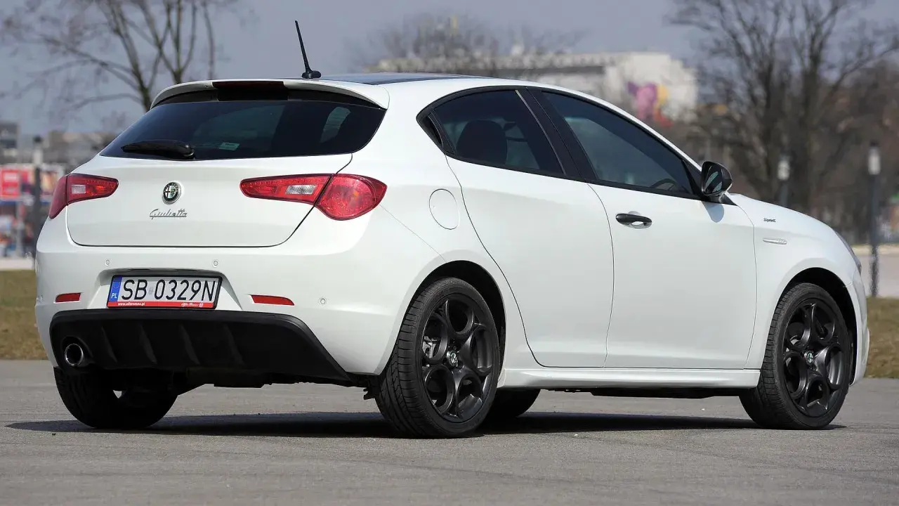 Biała Alfa Romeo Giulietta z czarnymi felgami stoi na asfalcie. Widoczny tył samochodu z tablicą rejestracyjną SB 0329N.