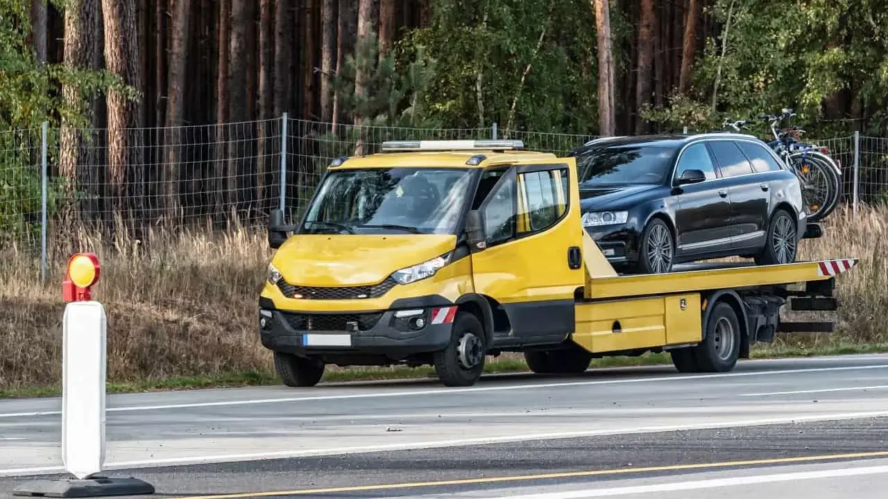 Żółta laweta z czarnym autem na pace, zaparkowana przy drodze. Obok stoi pachołek ostrzegawczy. To może być kara za parkowanie w miejscu niedozwolonym.