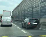 Korek na autostradzie. Samochody osobowe i ciężarowe stoją w rzędzie. W tle widać ekrany akustyczne. Jazda na suwak przepis to podstawa.