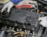 Mechanik w białych rękawiczkach sprawdza silnik, odkręcając korek wlewu oleju. Widać elementy rozrządu i pokrywę zaworów.