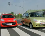 Czerwony i zielony samochód na pasach. Czy w terenie zabudowanym można wyprzedzać? Kierowcy zbliżają się do skrzyżowania.