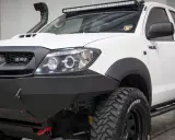 Biała Toyota Hilux z potężnym stalowym zderzakiem, wyciągarką i snorkel'em. Idealna do terenowych wypraw, gdzie przepisy nie ograniczają.