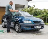 Mężczyzna myje niebieskie auto pod domem w upalny dzień. Wąż ogrodowy leży na podjeździe.