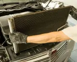 Mechanik w rękawicach wyjmuje duży, czarny skraplacz klimatyzacji z komory silnika samochodu.