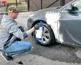 Mężczyzna w czapce z daszkiem i jeansach kuca przy samochodzie, rozpylając pianę na błotnik. Czy można naprawiać auto pod blokiem? Tak, jeśli to drobna naprawa lakieru.