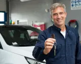 Uśmiechnięty mechanik w granatowym kombinezonie podaje kluczyki do białego samochodu. To symbol zakończenia prac i potwierdzenia, że co to homologacja jest już załatwiona.