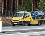 Żółta laweta z czarnym autem na pace, zaparkowana przy drodze. Obok stoi pachołek ostrzegawczy. To może być kara za parkowanie w miejscu niedozwolonym.