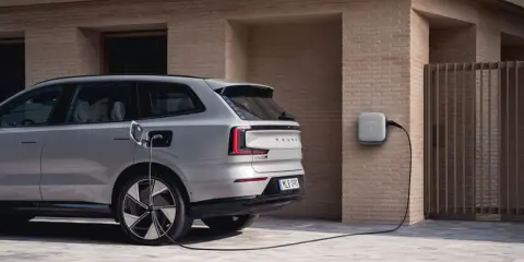 Srebrny, duży SUV Volvo EX90 podłączony do ładowarki ściennej. Nowoczesny design i ekologiczna energia.