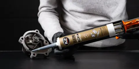 Ręce w czarnych rękawiczkach aplikują czarny silikon K2 na uszczelkę pompy wodnej.