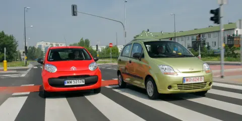Czerwony i zielony samochód na pasach. Czy w terenie zabudowanym można wyprzedzać? Kierowcy zbliżają się do skrzyżowania.