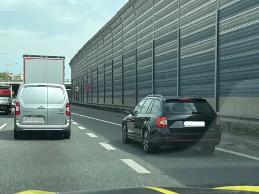 Korek na autostradzie. Samochody osobowe i ciężarowe stoją w rzędzie. W tle widać ekrany akustyczne. Jazda na suwak przepis to podstawa.