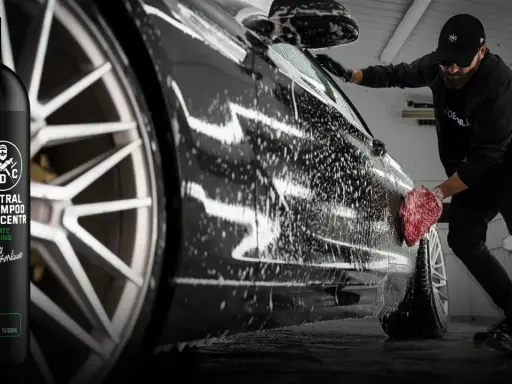 Mężczyzna myje czarne auto pianą, używając gąbki. Obok butelka szamponu SWAG.