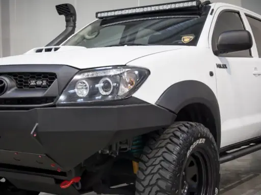 Biała Toyota Hilux z potężnym stalowym zderzakiem, wyciągarką i snorkel'em. Idealna do terenowych wypraw, gdzie przepisy nie ograniczają.