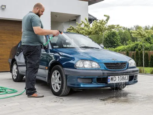 Mężczyzna myje niebieskie auto pod domem w upalny dzień. Wąż ogrodowy leży na podjeździe.