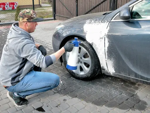 Mężczyzna w czapce z daszkiem i jeansach kuca przy samochodzie, rozpylając pianę na błotnik. Czy można naprawiać auto pod blokiem? Tak, jeśli to drobna naprawa lakieru.