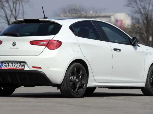 Biała Alfa Romeo Giulietta z czarnymi felgami stoi na asfalcie. Widoczny tył samochodu z tablicą rejestracyjną SB 0329N.