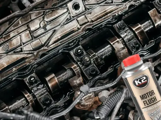 Widok wnętrza silnika z widocznymi wałkami rozrządu i śladami oleju. Obok butelka płukanki silnika K2 Motor Flush, sugerująca problemy po jej użyciu.