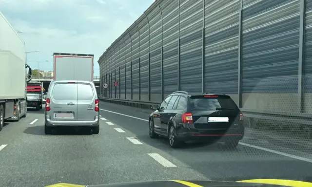 Korek na autostradzie. Samochody osobowe i ciężarowe stoją w rzędzie. W tle widać ekrany akustyczne. Jazda na suwak przepis to podstawa.
