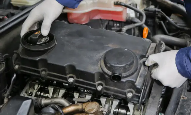 Mechanik w białych rękawiczkach sprawdza silnik, odkręcając korek wlewu oleju. Widać elementy rozrządu i pokrywę zaworów.