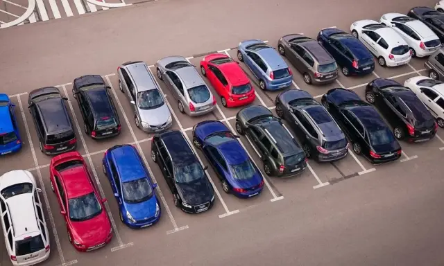 Rząd samochodów na parkingu, w tym czerwony, niebieski i srebrny. Jeden z nich czeka na odholowanie.