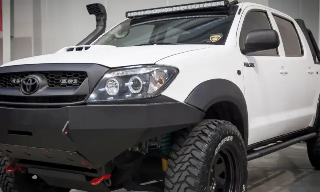 Biała Toyota Hilux z potężnym stalowym zderzakiem, wyciągarką i snorkel'em. Idealna do terenowych wypraw, gdzie przepisy nie ograniczają.
