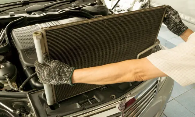 Mechanik w rękawicach wyjmuje duży, czarny skraplacz klimatyzacji z komory silnika samochodu.