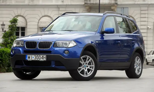 Niebieskie BMW E83, SUV, stoi przed budynkiem z łukami.