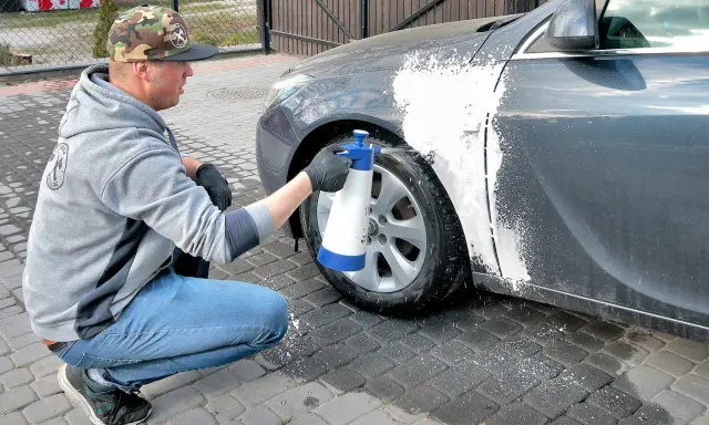 Mężczyzna w czapce z daszkiem i jeansach kuca przy samochodzie, rozpylając pianę na błotnik. Czy można naprawiać auto pod blokiem? Tak, jeśli to drobna naprawa lakieru.