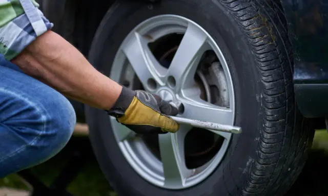 Mężczyzna w rękawiczkach dokonuje wymiany koła w samochodzie, używając klucza do odkręcenia śrub.