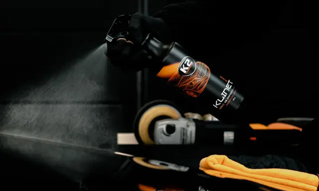 Rękawiczka w czarnej rękawiczce rozpyla produkt K2 KLINET na czarną powierzchnię, sprawdzając, czy aceton niszczy lakier samochodowy.