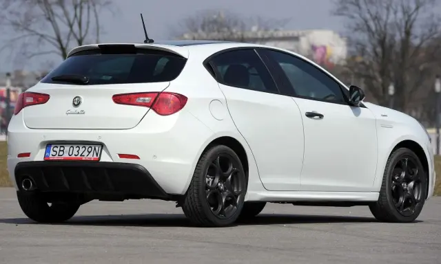 Biała Alfa Romeo Giulietta z czarnymi felgami stoi na asfalcie. Widoczny tył samochodu z tablicą rejestracyjną SB 0329N.