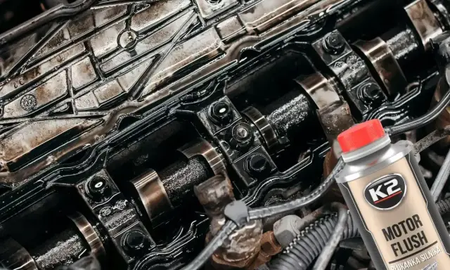 Widok wnętrza silnika z widocznymi wałkami rozrządu i śladami oleju. Obok butelka płukanki silnika K2 Motor Flush, sugerująca problemy po jej użyciu.