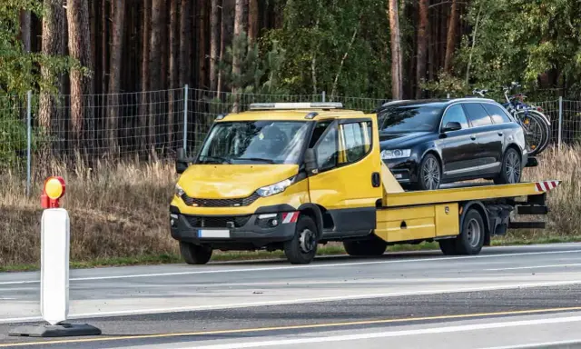 Żółta laweta z czarnym autem na pace, zaparkowana przy drodze. Obok stoi pachołek ostrzegawczy. To może być kara za parkowanie w miejscu niedozwolonym.