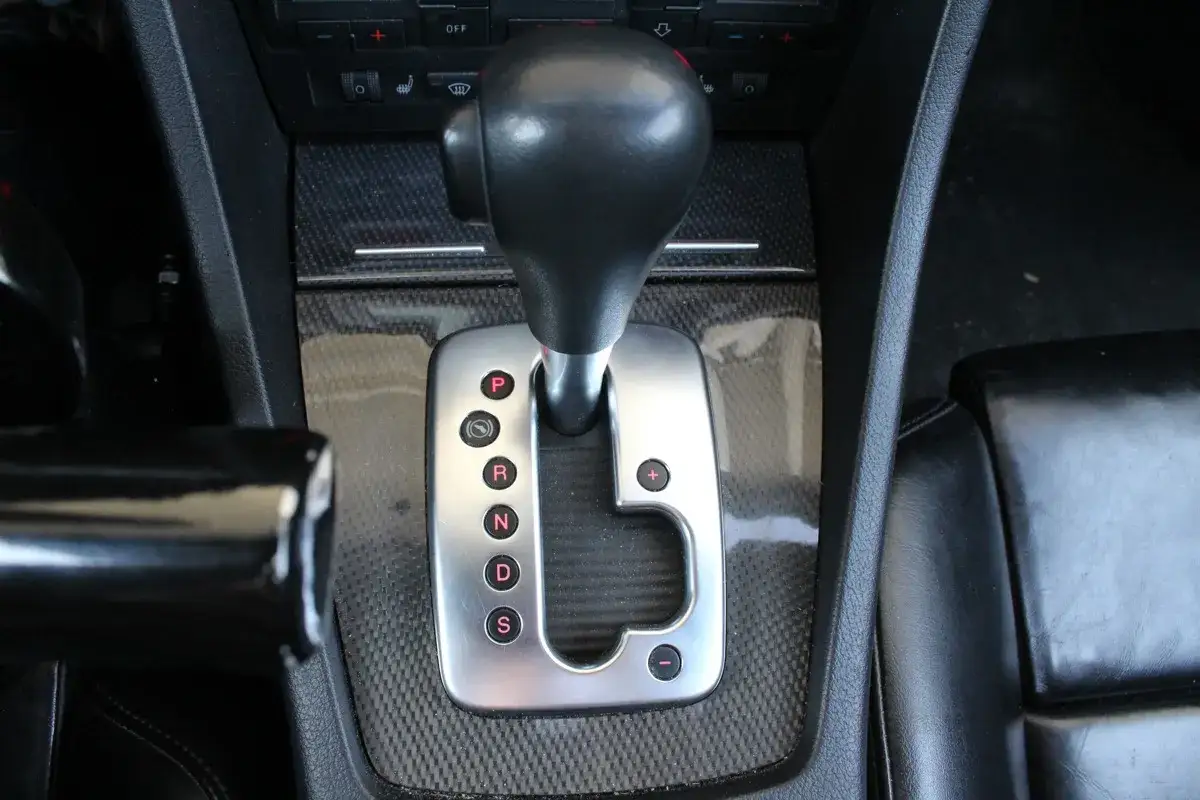 Dźwignia skrzyni tiptronic z oznaczeniami P, R, N, D, S. Widoczny fragment wnętrza samochodu z czarną tapicerką.
