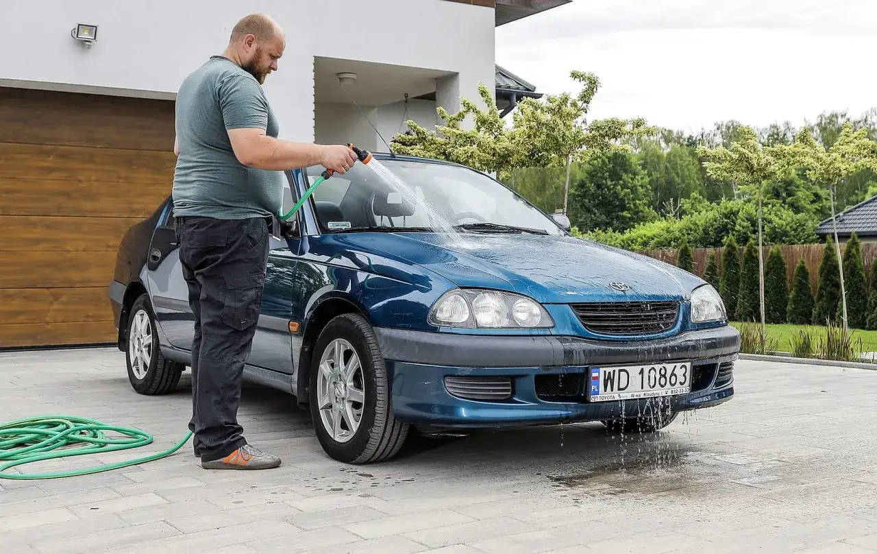 Mężczyzna myje niebieski samoch&oacute;d pod domem, używając węża ogrodowego. Woda spływa po masce i zderzaku.
