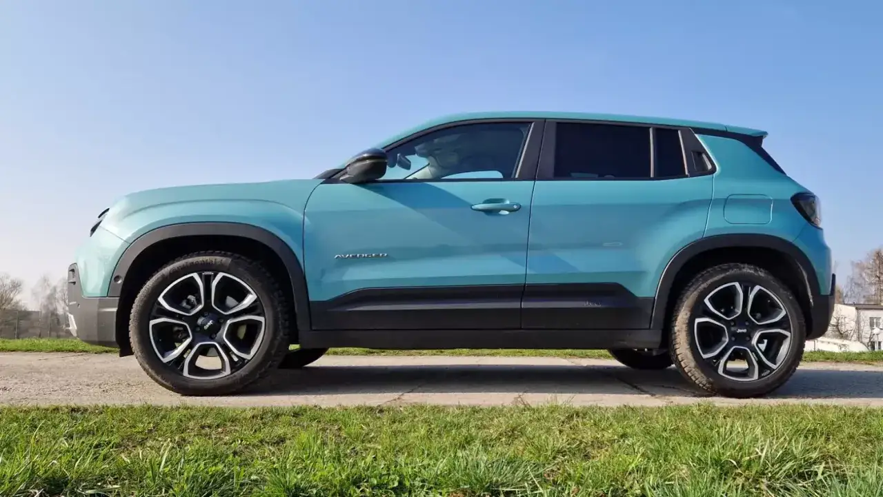 Turkusowy, duży SUV Jeep Avenger stoi na tle błękitnego nieba.