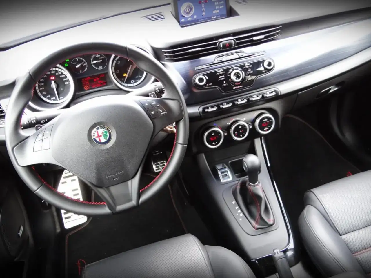 Wnętrze Alfa Romeo Giulietta: kierownica z logo, deska rozdzielcza z radiem i klimatyzacją, dźwignia zmiany bieg&oacute;w.