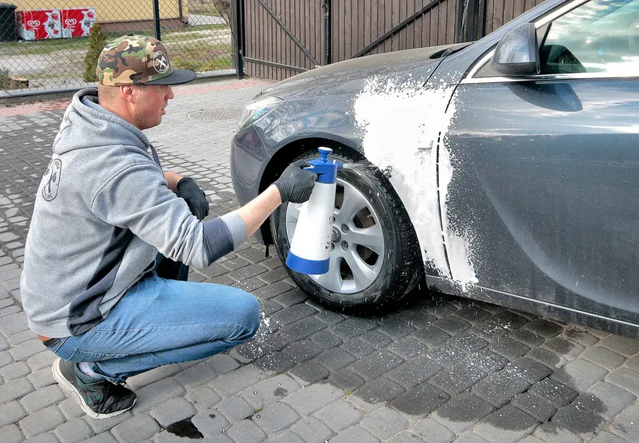 Mężczyzna w czapce z daszkiem i dżinsach kuca przy samochodzie, rozpylając pianę na błotnik. Czy można naprawiać auto pod blokiem? Tak, jeśli masz odpowiednie narzędzia.