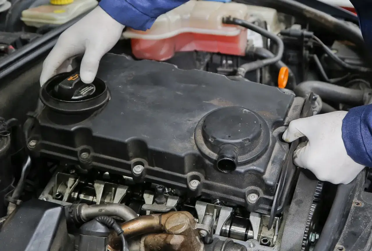 Mechanik w białych rękawiczkach sprawdza silnik, odkręcając korek wlewu oleju. Widać elementy układu rozrządu i przewody.