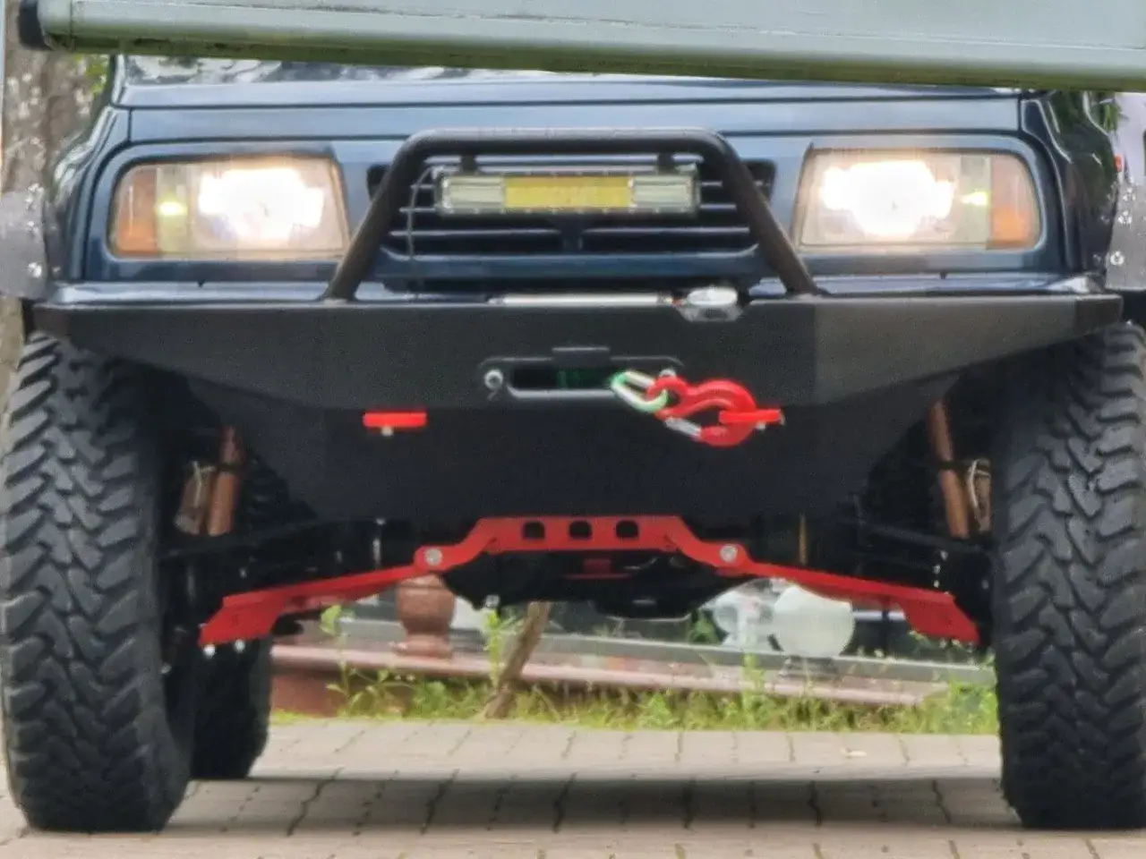 Ciemnoniebieski SUV z wyciągarką i mocnym zderzakiem. Widoczne czerwone elementy zawieszenia i opony terenowe. Idealny do jazdy w trudnym terenie, z napędem 4x4 dołączanym ręcznie.