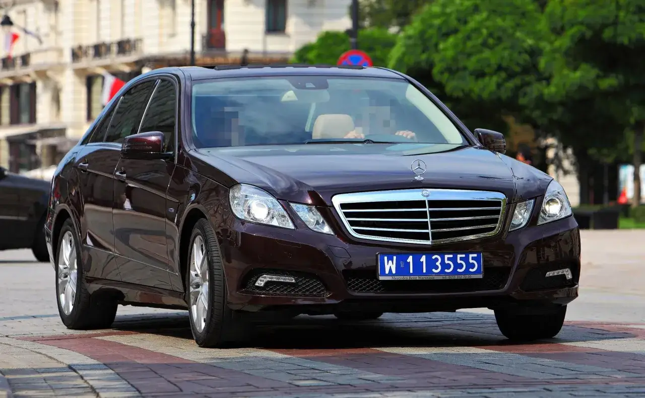 Ciemnobrązowy Mercedes z dyplomatycznymi tablicami rejestracyjnymi W 113555 na tle budynków i zieleni.