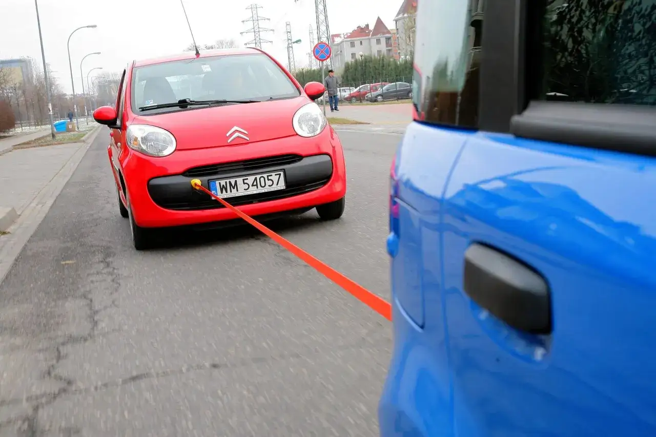 Czerwony Citroen jest holowany przez niebieski samoch&oacute;d. Pamiętaj o przepisach dotyczących holowania auta, by było bezpiecznie.